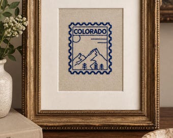 May include: A framed embroidery piece featuring a blue stitched design of a postage stamp with the word COLORADO, mountain peaks, a sun, and pine trees on a light beige fabric background, enclosed in an ornate metallic gold frame.