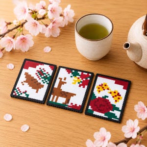 以下が含まれることがあります： ピクセルアートデザインの長方形コースター3枚。1枚目は茶色の鹿、2枚目は立っている鹿、3枚目は赤い花が描かれています。各コースターは黒い縁取りで、白い背景にカラフルな幾何学模様が施されています。