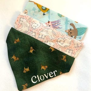 May include: Three triangular pet bandanas stacked together. The top one is light blue with a yellow submarine and mermaid cat print. The middle one is light pink with a white cat pattern. The bottom one is dark green with orange cats and the name Clover.