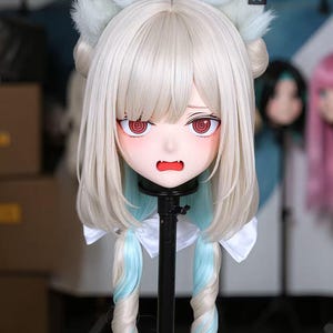 May include: A white and light blonde anime-style wig with large, fluffy, light blue ears. The wig features a detailed face with red eyes and a small open mouth. The hair is styled with light blue accents and long, twisted pigtails.