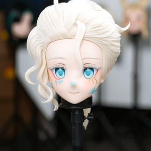 May include: A stylized doll head featuring pale skin, bright blue eyes, and voluminous white hair styled with a side-swept fringe. The face displays delicate blue and gold markings on the cheeks, a small blue nose detail, and a neutral expression.