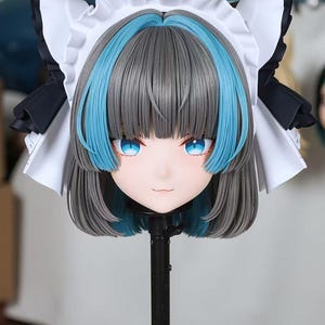 May include: A detailed close-up of a doll head with gray hair, blue highlights, and bright blue eyes. The doll head is wearing a white maid's headpiece with black accents. The face has a gentle expression and is mounted on a black stand.