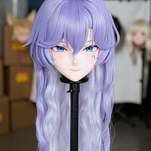 May include: A mask with a serene expression, featuring blue eyes and a lavender wig with long, flowing, white-tipped hair. The mask is mounted on a black stand, with a blurred background.