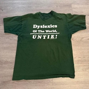 May include: A dark green t-shirt featuring the white text "Dyslexics Of The World, UNTIE!". The shirt has a crew neck and short sleeves. The text is centered on the front of the garment.