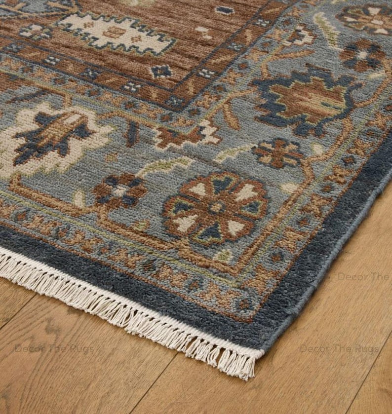 May include: Close-up of a rectangular rug with a detailed floral pattern. The rug features brown, blue, and beige tones. The border is a darker blue, and the rug has a fringe on one side. The rug is placed on a wooden floor.