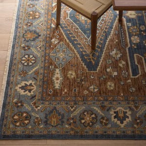 May include: A close-up of a patterned rug in brown and blue hues. The rug has a central brown section with geometric patterns and a blue border. A wooden chair and table leg are partially visible.