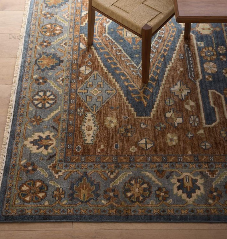 May include: A close-up of a patterned rug in brown and blue hues. The rug has a central brown section with geometric patterns and a blue border. A wooden chair and table leg are partially visible.