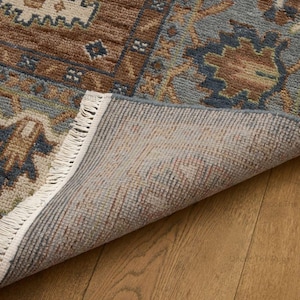 May include: Close-up of a patterned rug with a section folded back, showing the underside. The rug has a brown, blue, and beige design with a fringed edge. It rests on a wooden floor. The rug's design is intricate.