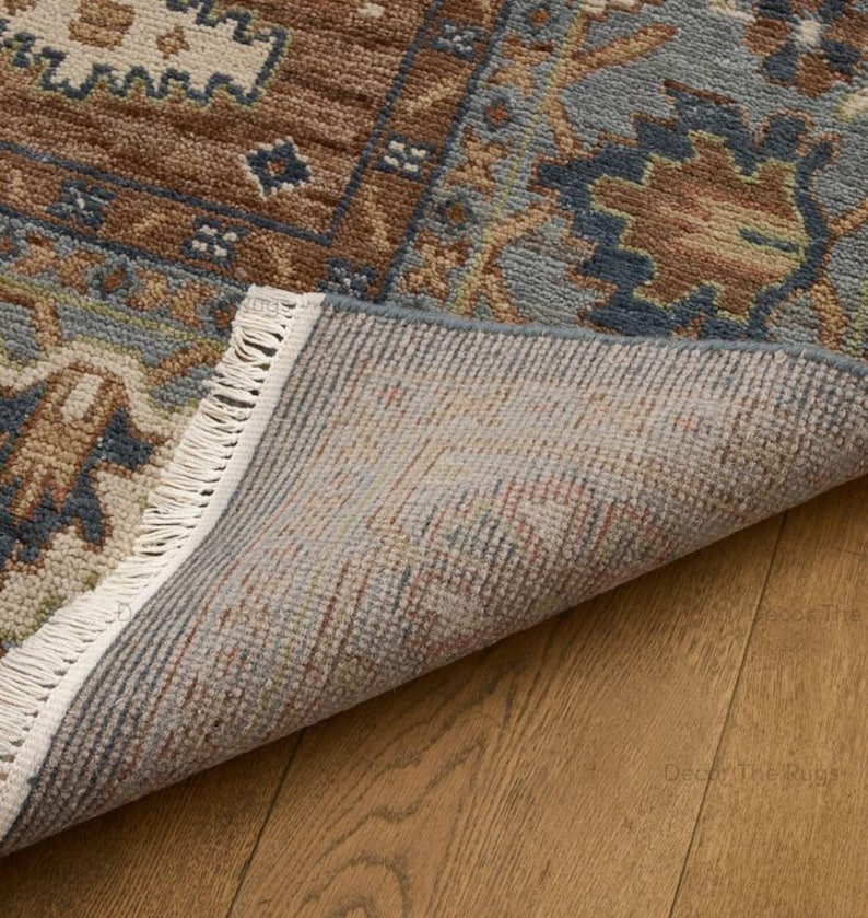 May include: Close-up of a patterned rug with a section folded back, showing the underside. The rug has a brown, blue, and beige design with a fringed edge. It rests on a wooden floor. The rug's design is intricate.