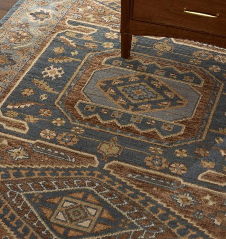 May include: A close-up of a blue and brown patterned rug with a geometric design. The rug features a central medallion and border with intricate floral and geometric motifs. A wooden cabinet leg is visible in the upper right corner.