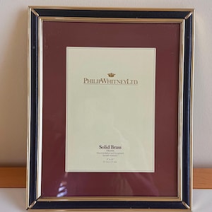May include: A rectangular picture frame with a black and gold border. It features a burgundy mount and a cream-colored interior with the text "PHILIP WHITNEY LTD." and "Solid Brass". The frame measures 20.3 cm x 25.4 cm.