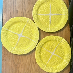 May include: Three round plates in a bright yellow color, each featuring a textured surface that mimics the appearance of a sliced lemon. The design includes a central white cross-like pattern representing the pith and segments of the fruit.