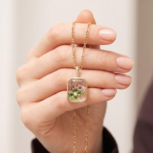 May include: Gold-toned necklace with a rectangular pendant. The pendant features a clear center with a green four-leaf clover design and the word "FUCK" in white. The necklace is held by a person.