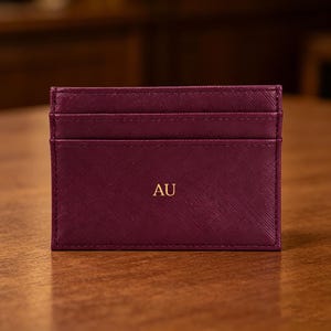 May include: A burgundy card holder with three card slots. The front of the holder has the gold letters "AU". The card holder is made of a textured material and is sitting on a wooden surface.