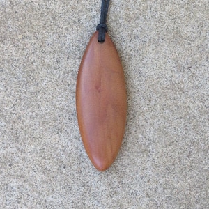 May include: A brown, oval-shaped pendant made of wood, suspended from a black cord. The pendant has a smooth, polished surface and a natural wood grain. The necklace is displayed against a textured, gray background.