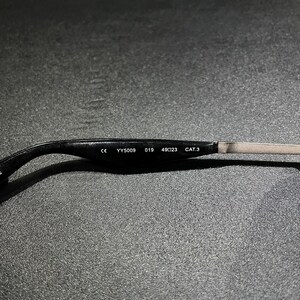 May include: The inner side of a black eyeglass temple arm featuring white printed text that reads CE YY5009 019 49 23 CAT.3. The arm transitions from a curved black plastic end piece to a thin, straight metallic segment on the right side.