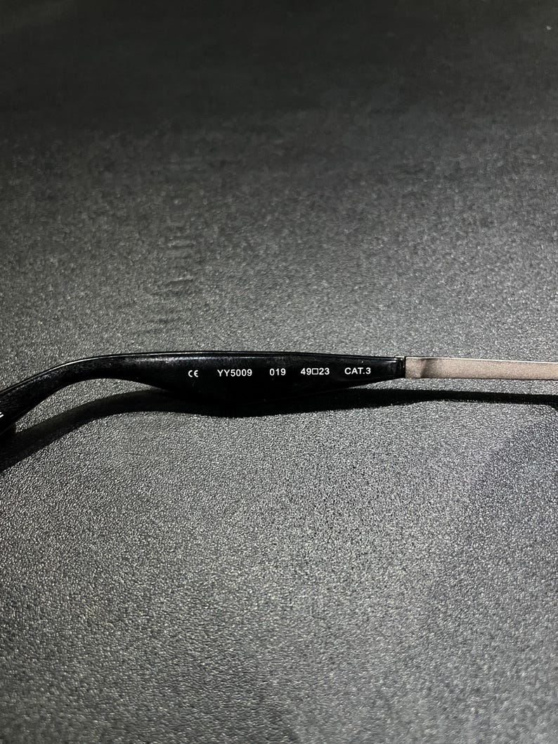 May include: The inner side of a black eyeglass temple arm featuring white printed text that reads CE YY5009 019 49 23 CAT.3. The arm transitions from a curved black plastic end piece to a thin, straight metallic segment on the right side.