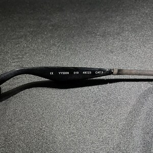 May include: The inner side of a black eyeglass temple arm featuring white printed text that reads CE YY5009 019 49 23 CAT.3. The arm transitions from a smooth black material to a thin metallic component on the right side of the frame.