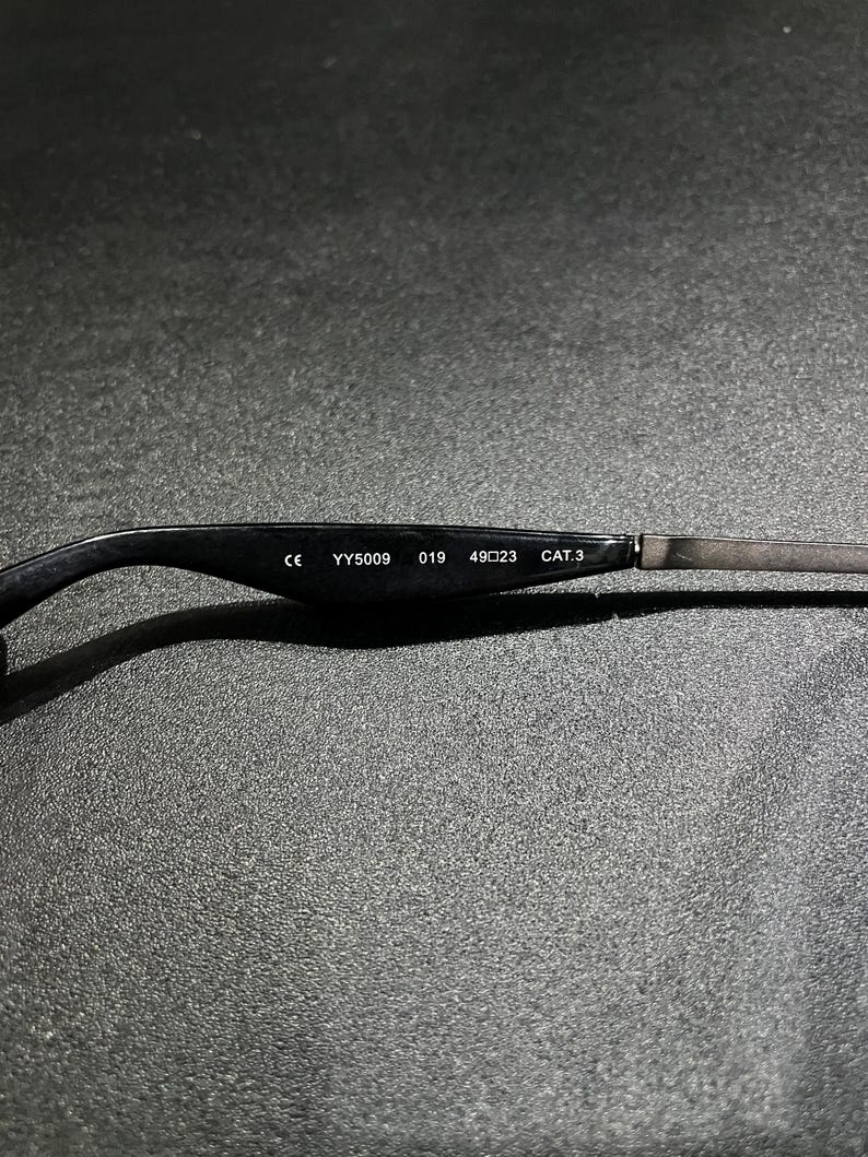 May include: The inner side of a black eyeglass temple arm featuring white printed text that reads CE YY5009 019 49 23 CAT.3. The arm transitions from a smooth black material to a thin metallic component on the right side of the frame.