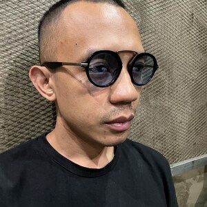 May include: Round sunglasses featuring a black frame with a distinct double bridge design and dark tinted lenses. The temples are thin and metallic, connecting to the circular rims. These eyewear pieces provide a classic look with a modern structural twist.
