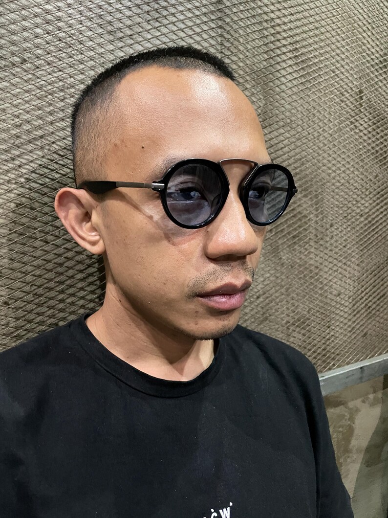 May include: Round sunglasses featuring a black frame with a distinct double bridge design and dark tinted lenses. The temples are thin and metallic, connecting to the circular rims. These eyewear pieces provide a classic look with a modern structural twist.