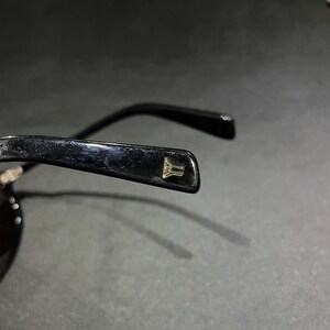 May include: A pair of black sunglasses featuring a glossy finish on the temple arm. A small, gold-colored metallic emblem shaped like a stylized letter Y is embedded into the end of the temple tip, providing a subtle contrast to the black frame.
