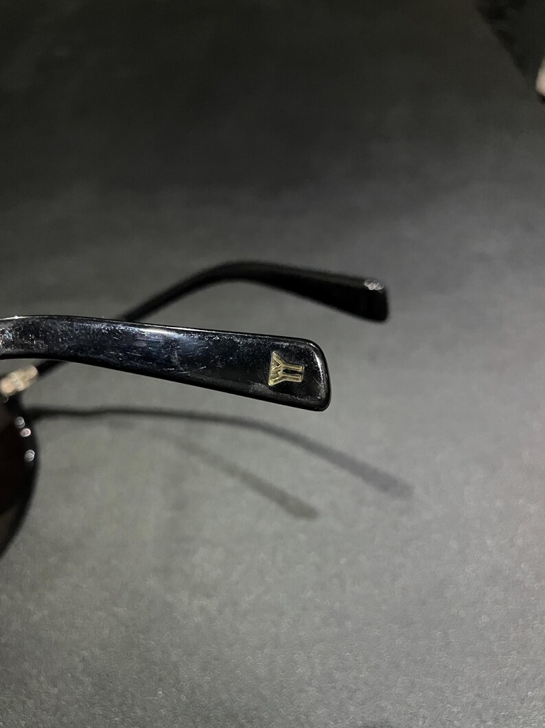 May include: A pair of black sunglasses featuring a glossy finish on the temple arm. A small, gold-colored metallic emblem shaped like a stylized letter Y is embedded into the end of the temple tip, providing a subtle contrast to the black frame.