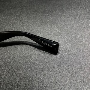 May include: A black glossy eyeglass temple arm featuring a signature script logo engraved on the side. The curved plastic frame component displays a smooth, reflective surface with a distinct, stylized handwritten brand name in white lettering.