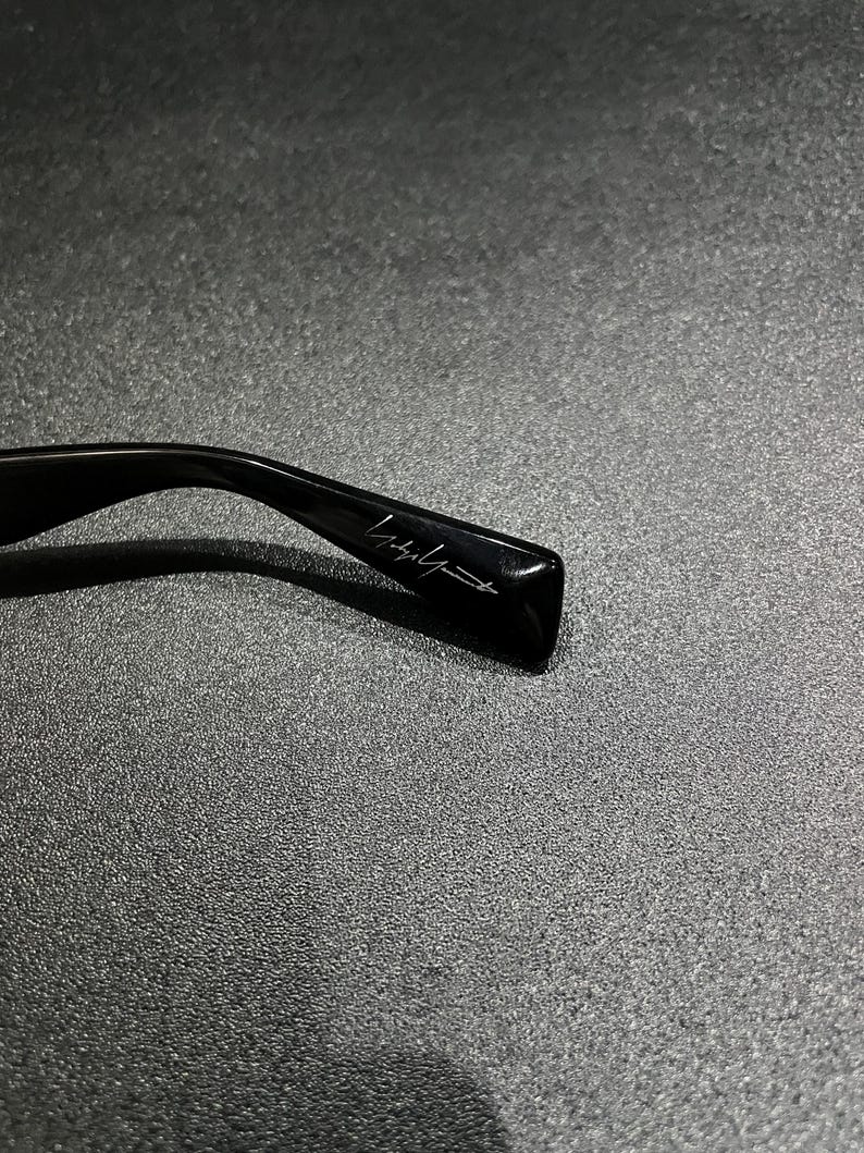 May include: A black glossy eyeglass temple arm featuring a signature script logo engraved on the side. The curved plastic frame component displays a smooth, reflective surface with a distinct, stylized handwritten brand name in white lettering.