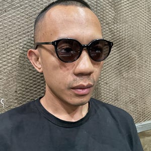 May include: Black-framed sunglasses with dark, round lenses and silver-toned rivets on the front corners. The frames feature a smooth, glossy finish and thin temples that extend along the sides of the head, providing a classic, rounded silhouette.