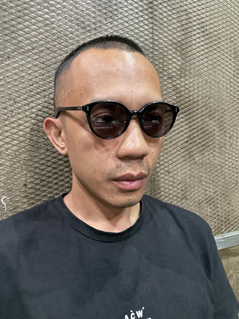 May include: Black-framed sunglasses with dark, round lenses and silver-toned rivets on the front corners. The frames feature a smooth, glossy finish and thin temples that extend along the sides of the head, providing a classic, rounded silhouette.