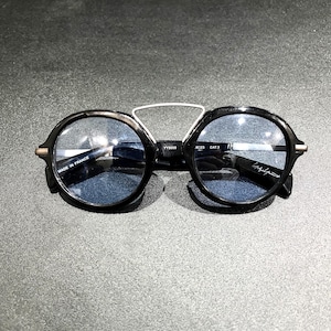 May include: Round sunglasses with black frames and blue-tinted lenses. A silver-toned metal bridge connects the top of the frames. The inner temple features the text "MADE IN FRANCE" and "YY5009", while the other lens displays a signature.