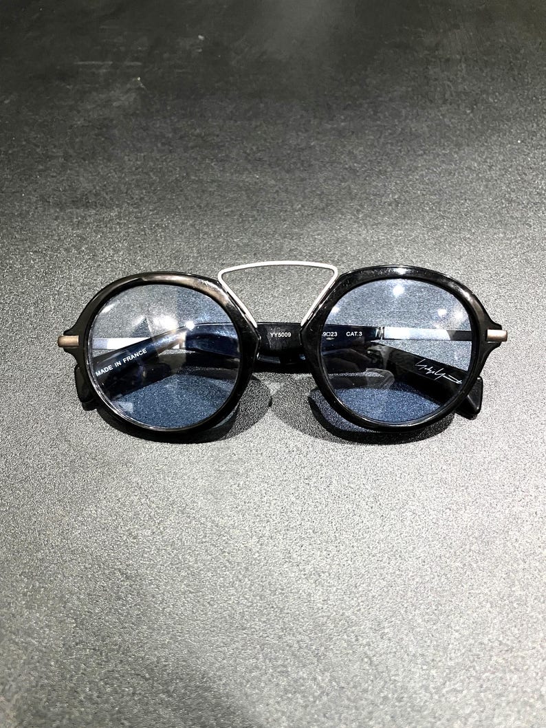 May include: Round sunglasses with black frames and blue-tinted lenses. A silver-toned metal bridge connects the top of the frames. The inner temple features the text "MADE IN FRANCE" and "YY5009", while the other lens displays a signature.