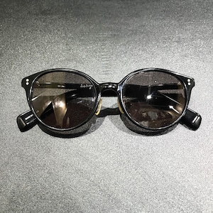 May include: Black sunglasses with round frames and dark tinted lenses. The frames feature two small silver-tone dots on each outer corner. The interior of the temple arm displays the text "MADE IN JAPAN" and a signature logo is visible on the lens.