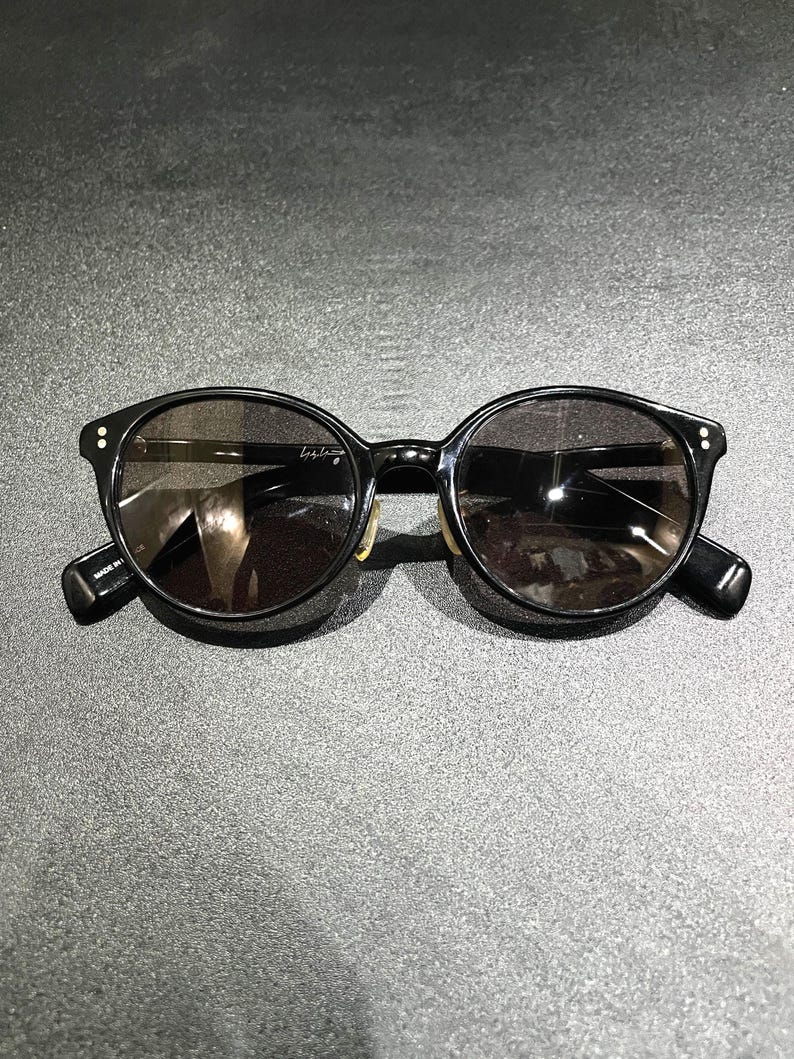 May include: Black sunglasses with round frames and dark tinted lenses. The frames feature two small silver-tone dots on each outer corner. The interior of the temple arm displays the text "MADE IN JAPAN" and a signature logo is visible on the lens.