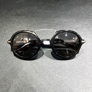 May include: Black round sunglasses featuring a distinctive silver-toned metal bridge connecting the two circular frames. The inner temple arm is clearly marked with the text MADE IN FRANCE and CAT 3, indicating the lens tint category for these glasses.