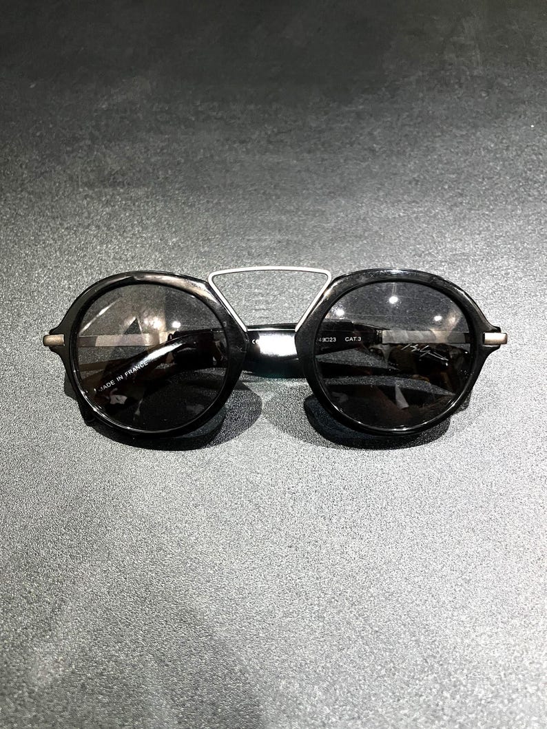 May include: Black round sunglasses featuring a distinctive silver-toned metal bridge connecting the two circular frames. The inner temple arm is clearly marked with the text MADE IN FRANCE and CAT 3, indicating the lens tint category for these glasses.