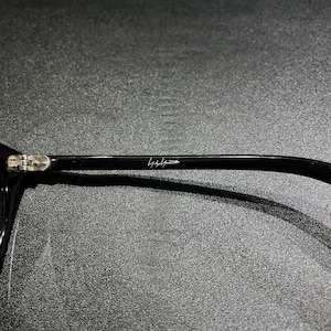 May include: A black eyeglass temple arm featuring a metallic hinge and a white signature logo printed on the side. The smooth, glossy black surface of the frame arm contrasts with the silver-toned metal hinge mechanism visible at the far left.