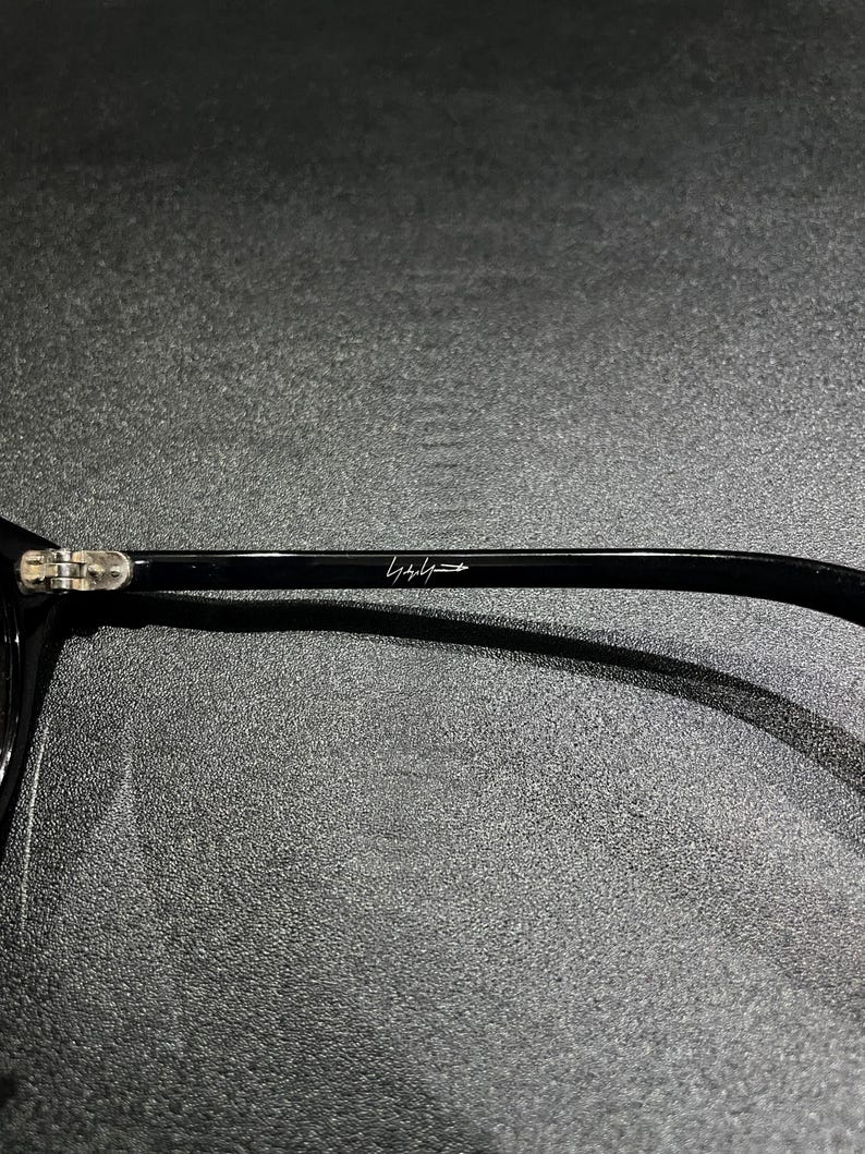 May include: A black eyeglass temple arm featuring a metallic hinge and a white signature logo printed on the side. The smooth, glossy black surface of the frame arm contrasts with the silver-toned metal hinge mechanism visible at the far left.