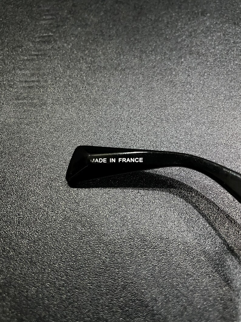 May include: A black temple arm of a pair of glasses featuring the text MADE IN FRANCE printed in white on the inner surface. The material has a smooth, glossy finish and a curved shape designed to rest behind the ear.