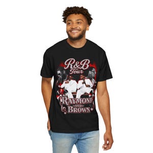 May include: A black short-sleeve t-shirt featuring a graphic print with the text R&B Tour and Raymond and Brown. The design includes two figures in white suits, musical notes, and red accents, set against a dark background with stylized lettering.