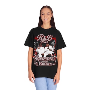 May include: A black short-sleeve t-shirt featuring a graphic print with the text R&B Tour Raymond and Brown. The design includes two figures in white suits, red circular accents, and stylized lettering in a glittery silver and red color scheme.