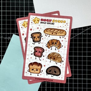 Könnte beinhalten: Ein Aufkleberbogen mit gezeichneten Brot-Charakteren mit wütenden Gesichtsausdrücken. Das Blatt enthält Illustrationen von Baguette, Croissant, Toast und Brotlaib mit dem Text "ANGY STARR ANGY BREAD" und "@ANGYSTARRSTUDIOS" auf einem rosa Hintergrund.