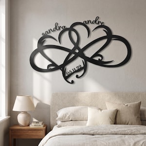 May include: A black metal wall art piece featuring two intertwined hearts forming an infinity symbol. The names "sandra" and "andre" are scripted above the hearts, and the date "05.11.22" is integrated into the lower section of the design.
