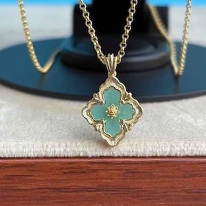 May include: A gold necklace with a four-leaf clover pendant. The pendant has a light green centre and a small gold flower. The pendant is framed by a detailed gold border, and the chain is also gold.