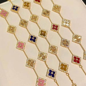 May include: Gold-coloured bracelets featuring square pendants with clover designs in pink, blue, red, and white. Each pendant is embellished with small, sparkling stones. The bracelets are presented on a white surface.