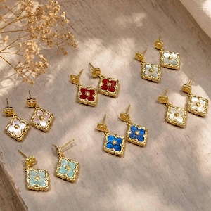 May include: Five pairs of gold-tone drop earrings, each featuring a four-leaf clover shape with a central star detail. The earrings come in various colors including red, white, light blue, and dark blue, all with a textured gold metal finish.