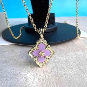 May include: A gold-coloured necklace with a pendant featuring a four-leaf clover design. The pendant has a light purple centre with a gold-coloured floral accent. The chain is a delicate link style.