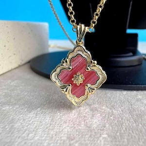 May include: A gold-tone necklace featuring a four-leaf clover pendant. The pendant has a red centre with a gold floral design. The necklace is displayed on a black stand against a light blue backdrop.