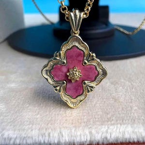 May include: A gold necklace with a pendant in the shape of a four-leaf clover, with a pink centre and gold filigree detailing. The pendant has a small gold flower in the centre. The chain is a gold cable chain.
